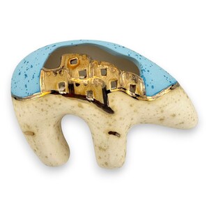 Könnte beinhalten: Eine bärenförmige Keramikfigur mit einem gesprenkelten cremefarbenen Körper und einem hellblau gesprenkelten Rücken. Der obere Teil zeigt ein goldenes Ausschnittdesign, das eine Reihe kleiner Gebäude oder Behausungen entlang des Rückens darstellt.
