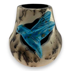 May include: A decorative vase with a unique design. The vase has a metallic finish with dark markings and a blue bird cutout. The bird has blue and gold accents. The interior of the vase is black.