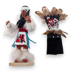 Puede incluir: Dos muñecas Kachina nativas americanas. Una muñeca está de pie y tiene una cinta blanca en la cabeza, cabello negro y un diseño pintado en rojo y blanco en la falda. La otra muñeca es un abanico de mano negro y rojo con plumas.