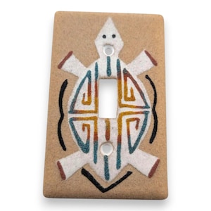 May include: Tan ceramic light switch cover featuring a white turtle design with teal, orange, and red accents. The turtle design is centered around the switch opening, with black curved lines on either side.