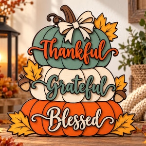 Può includere: Una decorazione a zucche sovrapposte composta da tre zucche impilate nei colori verde acqua, crema e arancione. Le zucche sono decorate con un fiocco crema, foglie d'acero e ghiande. Il testo Thankful, Grateful, Blessed è scritto in corsivo.