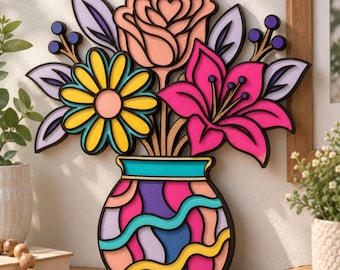 May include: A layered wooden wall art piece featuring a bouquet of flowers in a vase. The arrangement includes a peach rose, a yellow daisy, and a bright pink lily with purple and lavender leaves. The vase has a colorful wavy pattern in pink, purple, and teal.