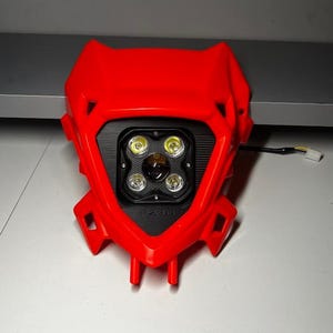 May include: A red motorcycle headlight assembly with a black center section. The headlight features four bright LED lights and the text "FZ3D" at the bottom. A wire with a connector extends from the back of the assembly.