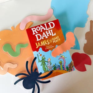 Puede incluir: Un libro infantil titulado "James y el melocotón gigante" de Roald Dahl, ilustrado por Quentin Blake. El libro está rodeado de formas recortadas, incluyendo una silueta azul, una araña y un melocotón.