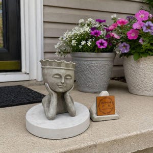May include: A gray concrete planter shaped like a serene face with a crown, resting on a round base. The planter is surrounded by potted flowers in shades of pink, purple, and white, creating a tranquil outdoor scene.