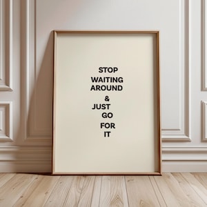 Könnte beinhalten: Ein gerahmter Druck mit dem Text "STOP WAITING AROUND & JUST GO FOR IT" in schwarzer Schrift. Der Druck hat einen cremefarbenen Hintergrund und einen Holzrahmen. Der Druck lehnt an einer weißen Wand und einem Holzboden.