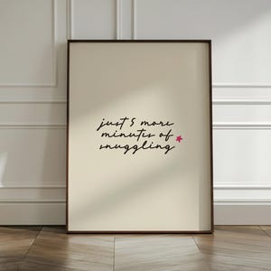 May include: Framed art print with the text "just 5 more minutes of snuggling" in a handwritten style, with a small pink star. The print has a cream-colored background and a dark brown frame. The artwork is displayed on a wooden floor.