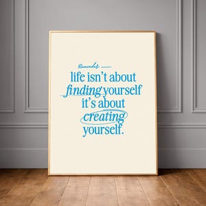 Puede incluir: Impresión enmarcada con un borde dorado. La obra de arte presenta la cita "life isn't about finding yourself it's about creating yourself" en escritura azul sobre un fondo crema. La impresión se exhibe sobre un suelo de madera.