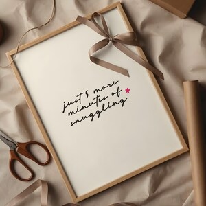 Könnte beinhalten: Ein gerahmter Druck mit dem Text "just 5 more minutes of snuggling" in schwarzer Schrift, akzentuiert mit einem rosa Stern. Oben ist eine braune Schleife gebunden. Schere und Geschenkpapier sind ebenfalls zu sehen.