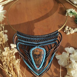 May include: A handcrafted macrame necklace featuring a large, teardrop-shaped stone pendant with brown, orange, and black hues. The pendant is framed by turquoise and black macrame, attached to a gold-tone metal collar.