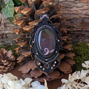 Peut inclure: Un collier pendentif en macramé avec une pierre ovale sombre aux inclusions rougeâtres, entourée d'un tressage complexe en cordon noir, d'accents en fil violet clair et de petites perles métalliques argentées, sur un cordon noir assorti.