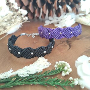 Peut inclure: Deux bracelets en macramé. L'un est noir avec des perles argentées, l'autre est violet avec des perles dorées. Les bracelets sont présentés sur une surface en bois avec des éléments décoratifs en arrière-plan. Les bracelets ont un motif tissé.