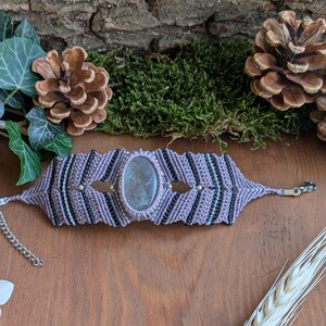 Peut inclure: Un bracelet en macramé avec une pierre ovale translucide grise au centre, sertie dans un motif de cordons tressés lavande et noirs. Le bracelet comprend une chaînette d'extension en métal argenté et un fermoir mousqueton réglable.