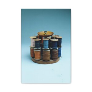 May include: A circular wooden thread rack featuring a central brass knob and multiple vertical spindles. The rack holds various wooden spools of sewing thread in shades of blue, brown, orange, and cream, arranged in a compact, organized display.