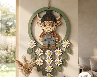 May include: A circular wooden wall hanging featuring a layered Highland cow character wearing a dark gray beanie and denim overalls. The design is framed by a green ring and decorated with white daisy flowers and green leaves hanging below.