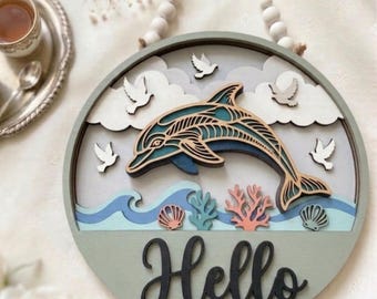 May include: A round, layered wooden wall hanging with a dolphin design. The dolphin is teal and blue, with waves, clouds, and the word "Hello" in black script. Includes a rope hanger and white wooden beads.