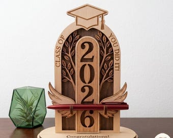 May include: A wooden graduation pen holder with the year 2026, a graduation cap, and the words "Class of" and "You did it!". The holder has a pen and the word "Congratulations!" at the base. The pen is red.