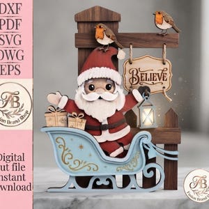 Puede incluir: Una artesanía de madera en capas que muestra a Santa Claus en un trineo azul claro con dos cajas de regalo. Un letrero colgado de un poste de madera oscura dice Believe, con dos pájaros pequeños posados encima. Incluye texto de archivos.