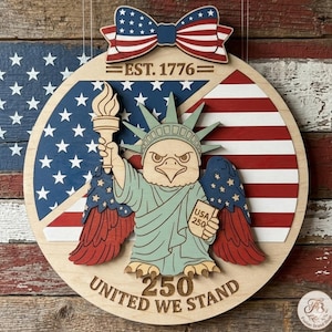 Puede incluir: Adorno de pared patriótico de madera con una Estatua de la Libertad estilizada con cabeza de águila, sosteniendo una antorcha y un letrero que dice "USA 250". El diseño incorpora la bandera estadounidense, el texto "EST. 1776" y "UNITED WE STAND".