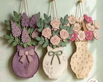 May include: Three wooden wall hangings featuring floral arrangements in vases. The left vase is purple with a bow and lavender, the center is cream with roses and a green bow, and the right is light wood with pink flowers and heart patterns.