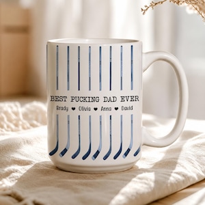 May include: A white ceramic mug featuring a repeating pattern of blue hockey sticks. The text "BEST PUCKING DAD EVER" is printed in black, followed by the names "Brady", "Olivia", "Anna", and "David" separated by small black heart icons.