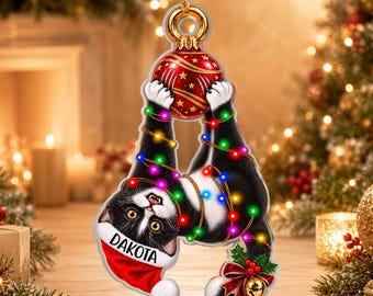 Könnte beinhalten: Ein flacher Acryl-Weihnachtsanhänger mit einer schwarz-weißen Katze, die eine rote Weihnachtsmütze mit dem Namen DAKOTA trägt. Die Katze ist in bunte Lichterketten verwickelt und hält eine rote, gemusterte Weihnachtskugel mit goldener Öse.