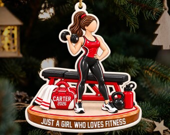 Könnte beinhalten: Ein hängendes Ornament mit einer Figur in rotem Tanktop und schwarzer Leggings, die eine Hantel hält. Die Figur steht neben einer roten Hantelbank, einer roten Sporttasche mit der Aufschrift CARTER 2026, einer Kettlebell und einer Trinkflasche. Text: JUST A GIRL WHO LOVES FITNESS.