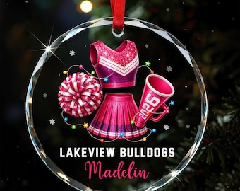 Könnte beinhalten: Ein rundes, klares Glasornament an einem roten Band mit einem glitzernden pinken Cheerleader-Outfit, einem Pompon und einem Megafon mit der Aufschrift 2026. Darunter steht LAKEVIEW BULLDOGS und der Name Madelin in pinker Schreibschrift.