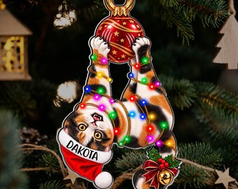 Könnte beinhalten: Ein hängender Weihnachtsbaumschmuck in Form einer Glückskatze mit einer roten Weihnachtsmütze, auf deren Rand der Name DAKOTA steht. Die Katze ist in bunte Lichterketten verwickelt und hält eine rote Weihnachtskugel sowie eine kleine Glocke.