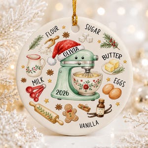 May include: A round white ceramic ornament featuring a mint green stand mixer wearing a Santa hat with the name Olivia and the year 2026. Surrounding the mixer are illustrations of baking ingredients labeled flour, sugar, butter, eggs, milk, and vanilla.