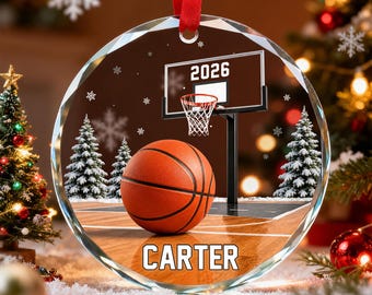 Könnte beinhalten: Ein runder, klarer Weihnachtsbaumschmuck aus Glas mit einem Basketball-Motiv, einem Korb mit der Jahreszahl 2026 und dem Namen CARTER in Weiß. Das Design zeigt verschneite Tannen und Schneeflocken vor einem dunklen Hintergrund.