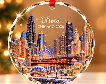 Könnte beinhalten: Ein rundes, klares Glasornament mit einem aufgedruckten nächtlichen Chicago-Stadtbild inklusive Schnee, Riesenrad und der Cloud Gate Skulptur. Der Text "Olivia" und "CHICAGO 2026" ist in Weiß aufgedruckt. Ein rotes Band ist oben befestigt.