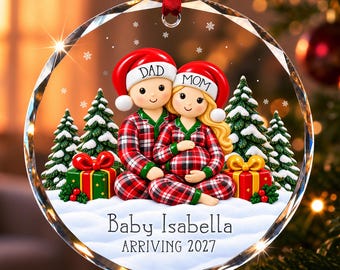 Könnte beinhalten: Ein runder, klarer Glas-Weihnachtsanhänger mit einem Paar in rot-karierten Schlafanzügen und Weihnachtsmützen mit der Aufschrift DAD und MOM. Die Szene zeigt Schnee, Tannenbäume und Geschenke. Text: Baby Isabella ARRIVING 2027.