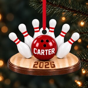 May include: A bowling-themed Christmas ornament featuring a red bowling ball with the name CARTER printed in white, surrounded by six white bowling pins with red stripes. The base is a wooden bowling lane with the year 2026 printed on the front.