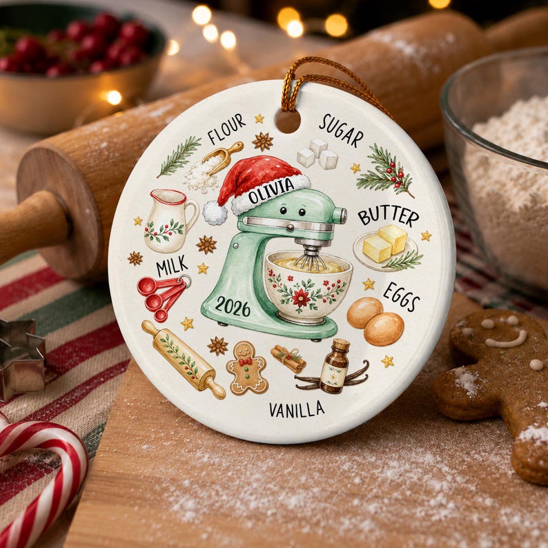 May include: A round white ceramic ornament featuring a light green stand mixer wearing a Santa hat with the name OLIVIA and the year 2026 printed on it. The design includes illustrations of baking ingredients labeled FLOUR, SUGAR, BUTTER, EGGS, MILK, and VANILLA.