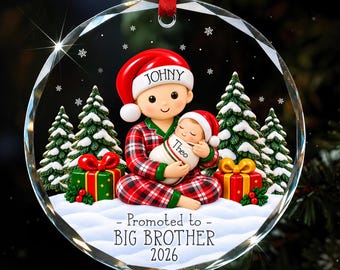 Könnte beinhalten: Ein runder, klarer Weihnachtsbaumschmuck aus Glas mit einem Kind im rot karierten Schlafanzug und Weihnachtsmannmütze namens Johny, das ein Baby namens Theo hält. Der Text lautet Promoted to BIG BROTHER 2026 vor verschneiten Bäumen.