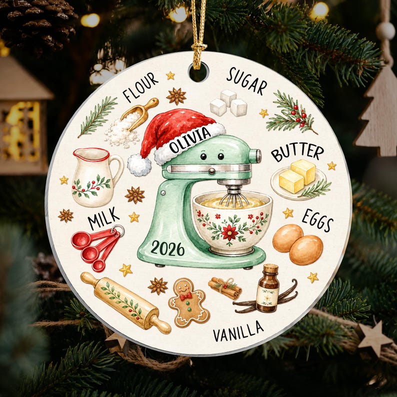 May include: A round ceramic ornament featuring a light green stand mixer wearing a Santa hat, labeled with the name Olivia and the year 2026. The design includes illustrations of baking ingredients like flour, sugar, butter, milk, eggs, and vanilla.