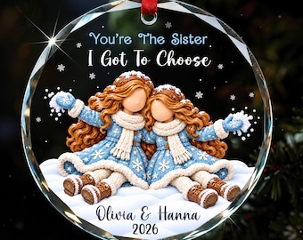 Könnte beinhalten: Ein runder Weihnachtsbaumschmuck aus Glas mit zwei Figuren mit langem braunem Haar, die blaue Mäntel, weiße Schals und braune Stiefel tragen. Der Text lautet: You're The Sister I Got To Choose, Olivia & Hanna, 2026. Die Figuren sitzen im Schnee.