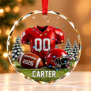 May include: A round, clear glass ornament featuring a red football jersey with the number 00, a brown football with the text 2026, and a red football helmet. The design includes snowy pine trees, colorful string lights, and the name CARTER.