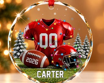 Könnte beinhalten: Ein rundes, klares Glasornament mit einem roten Football-Trikot mit der Nummer 00, einem braunen Football mit dem Text 2026 und einem roten Football-Helm. Das Design zeigt verschneite Tannenbäume, bunte Lichterketten und den Namen CARTER.