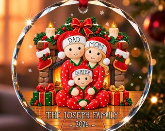 Könnte beinhalten: Ein runder Weihnachtsbaumschmuck aus Glas mit einer dreiköpfigen Familie in rot-gepunkteten Schlafanzügen und Weihnachtsmützen vor einem Kamin. Die Mützen tragen die Aufschriften DAD, MOM und QUATER. Unten steht THE JOSEPH FAMILY und 2026.