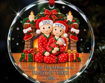 Könnte beinhalten: Ein runder Weihnachtsbaumschmuck aus Glas mit einer dreiköpfigen Familie in rot-gepunkteten Schlafanzügen vor einem Kamin. Die Figuren tragen Weihnachtsmützen mit der Aufschrift DAD, MOM und baby. Text: Our 1st Christmas as a FAMILY OF THREE - 2026.