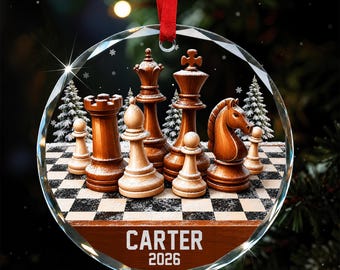 Könnte beinhalten: Ein runder Glasanhänger mit einem Schachspiel aus Holzfiguren auf einem Schachbrett, akzentuiert durch verschneite Tannenbäume. Der Anhänger hat ein rotes Band zum Aufhängen und zeigt den Text CARTER 2026 in Weiß auf einem braunen Sockel.