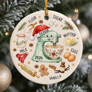May include: A round ceramic ornament featuring a light green stand mixer wearing a Santa hat with the name Olivia and the year 2026 printed on it. Surrounding the mixer are illustrations of baking ingredients labeled flour, sugar, butter, eggs, milk, and vanilla.