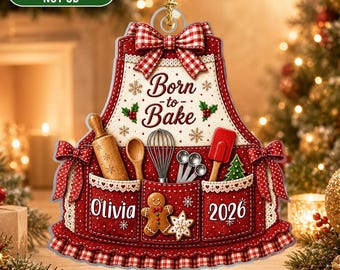 Könnte beinhalten: Ein flaches 2D-Acryl-Ornament in Schürzenform mit einer rot-weiß karierten Schleife, verziert mit dem Text Born to Bake, einem Nudelholz, Schneebesen, Messlöffeln, Teigschaber, einem Lebkuchenmann, dem Namen Olivia und der Jahreszahl 2026.