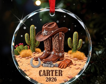 Könnte beinhalten: Ein rundes Glasornament mit rotem Band, das einen braunen Cowboyhut, Lederstiefel, ein Hufeisen, Sporen und Handschuhe in einer Wüstenlandschaft mit Kakteen zeigt. Der Text CARTER und 2026 ist unten in der Wüstenszene in Braun aufgedruckt.