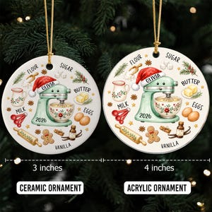 May include: Two round ornaments featuring a light green stand mixer with a Santa hat, the name Olivia, and the year 2026. Illustrations include flour, sugar, butter, eggs, milk, vanilla, and a gingerbread man. One is a 3-inch ceramic ornament, the other a 4-inch acrylic ornament.