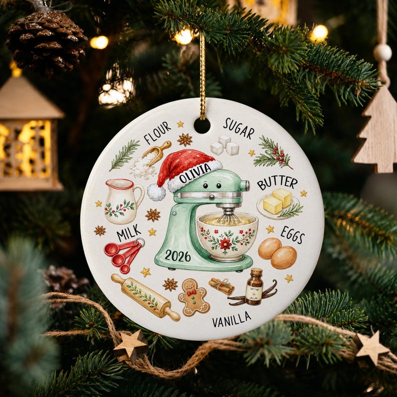 May include: A round white ceramic ornament featuring a light green stand mixer wearing a Santa hat with the name Olivia and the year 2026. Illustrations of baking ingredients like flour, sugar, butter, eggs, milk, and vanilla surround the mixer.