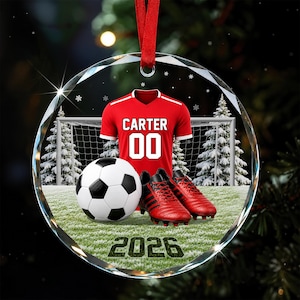 May include: A round glass ornament featuring a red soccer jersey with the name CARTER and number 00, a black and white soccer ball, and red soccer cleats. The background shows a soccer goal on a field with snowy trees and the year 2026.