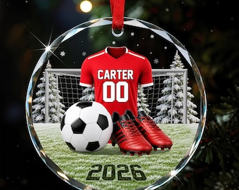 Könnte beinhalten: Ein rundes Glasornament mit einem roten Fußballtrikot mit dem Namen CARTER und der Nummer 00, einem schwarz-weißen Fußball und roten Fußballschuhen. Der Hintergrund zeigt ein Fußballtor auf einem Feld mit verschneiten Bäumen und dem Jahr 2026.